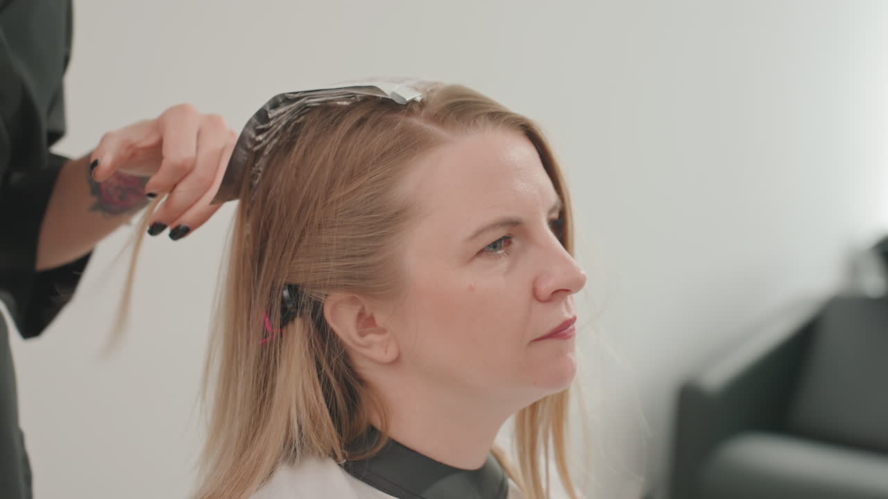 Mulher no salão fazendo luzes com papel alumínio. Cabeleireiro separa o cabelo e aplica papel alumínio e coloração na raiz com precisão, cliente sentada usando uma capa, interior moderno e bem iluminado, close-up nas mãos, clipes