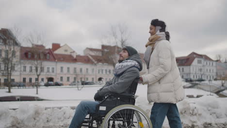 side view of happy muslim woman taking her disabled friend in wheelchair on a walk around the city in winter 2