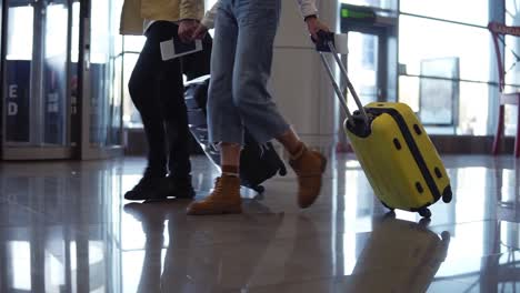 Cropped-front-footage-of-couple-travelers-are-carrying-their-luggage-and-holding-hands.-Walking-over-lounge-of-terminal.-People