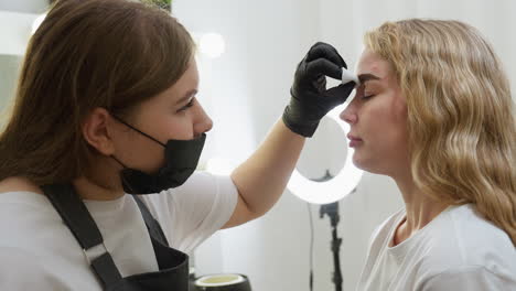 beautician wearing black gloves gently cleans eyebrow of relaxed client with cotton pad in well-lit beauty salon, with ring light and grooming tools visible in background, showcasing skincare session