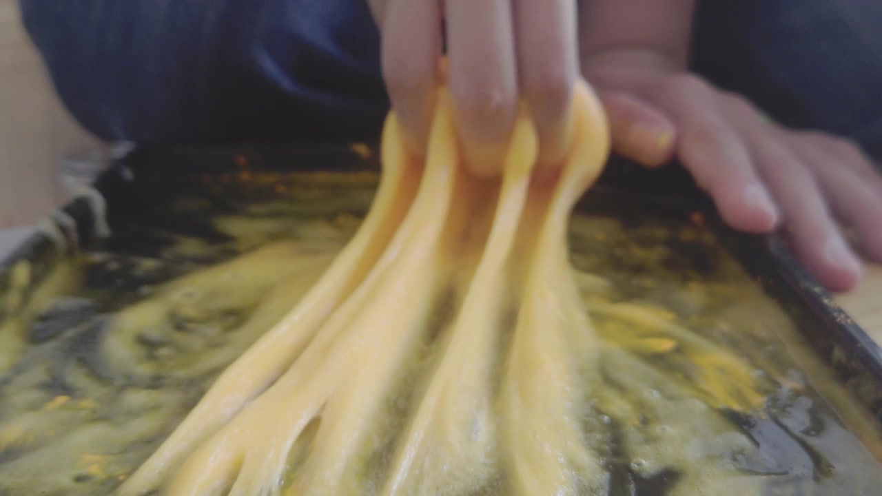 Uma foto em close-up de baixo ângulo mostra os dedos de uma criança levantando uma massa espessa e elástica de slime amarelo brilhante de uma bandeja de mistura escura, formando fios pegajosos verticais distintos durante uma experiência científica caseira.
