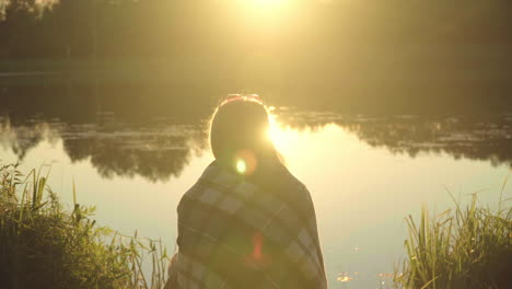 Lonely-girl-in-plaid-sitting-on-river-bank-in-rays-of-departing-sun.-Lonely-woman
