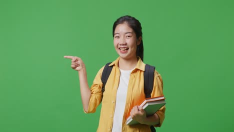 asian woman student with a backpack and some books smiling and pointing to side while standing in the green screen background studio