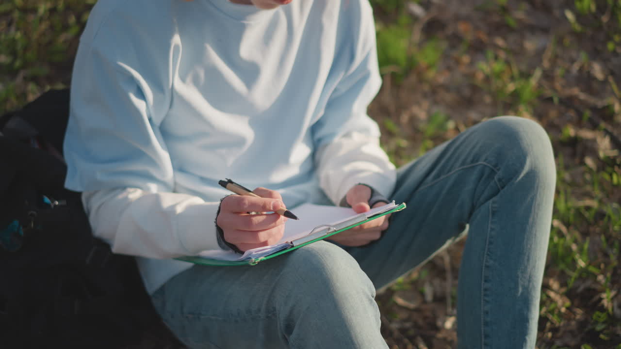 Студент в парке изучает конспекты: прилежный студент усердно записывает информацию в спокойной обстановке парка, молодой учащийся задумчиво делает заметки на открытом воздухе.
