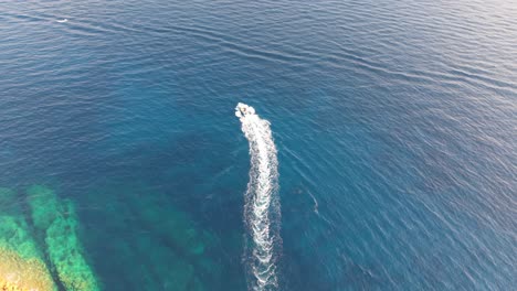 Drone video of a small speedboat creating a white trail as it passes by a rugged rocky coastline. The deep blue sea contrasts beautifully with the turquoise shallows near the shore