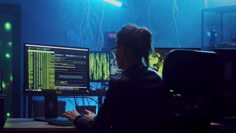 Rear-Of-The-Young-Girl-Hacker-In-Glasses-Working-At-The-Computer-At-Night-And-Hacking-A-Program-While-Her-Male-Colleague-In-A-Hood-Walking-The-Room