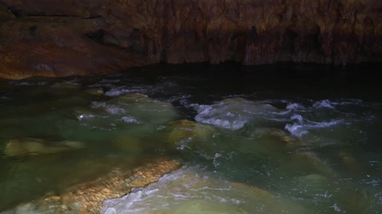 Clear emerald stream flows over textured rock formations inside a dimly lit cavern. Smooth handheld tracking shot follows the current revealing limestone walls and shallow pools.