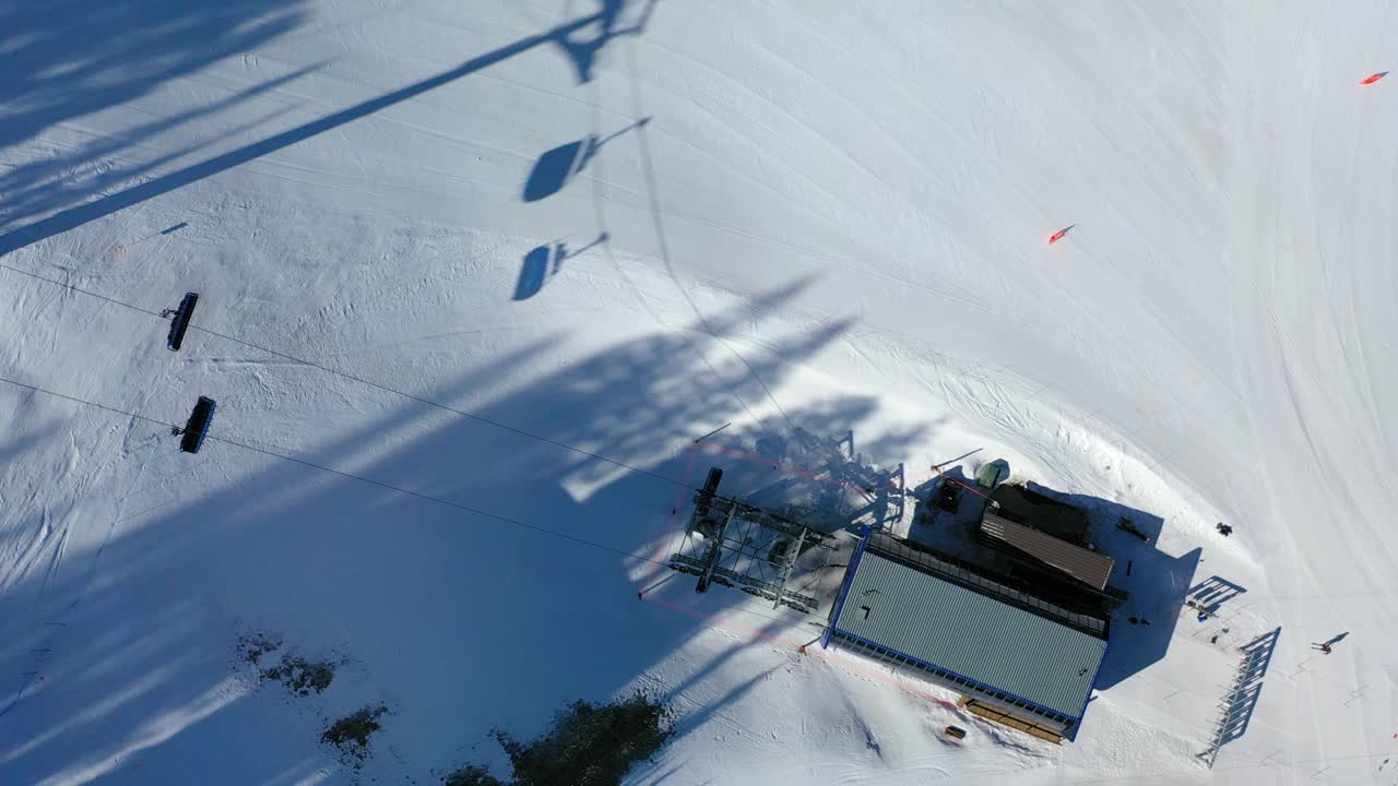 Drone captures an overhead view of skiers riding a snowy mountain chair lift, with scenic trails, tall evergreens, and distant white-capped peaks creating a perfect winter backdrop