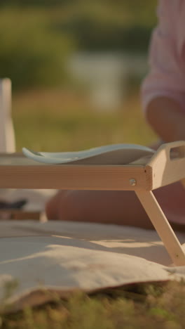mujer sentada al aire libre en una alfombra colocando un plato en una mesa de picnic con flores en una canasta tejida a su lado, está vestida con ropa casual, disfrutando de un picnic pacífico en una zona de hierba soleada