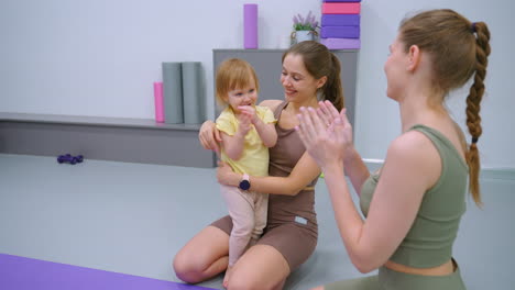 joy fills the gym as a mother holds her delighted daughter, both clapping hands with a friend nearby, surrounded by fitness equipment like dumbbells and foam rollers