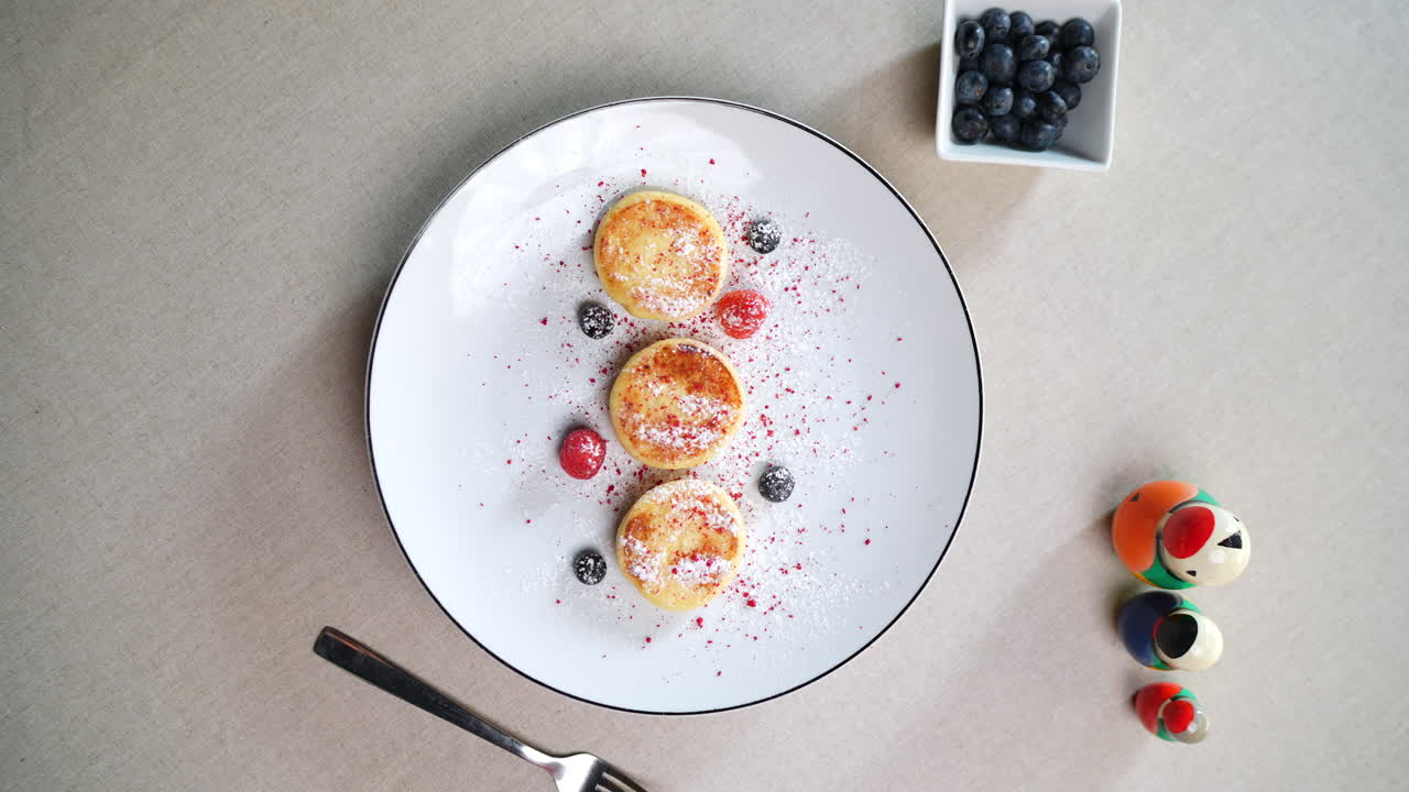 Traditional Eastern European cheese pancakes, syrniki, served on a white plate, garnished with berries and powdered sugar, alongside matryoshka dolls