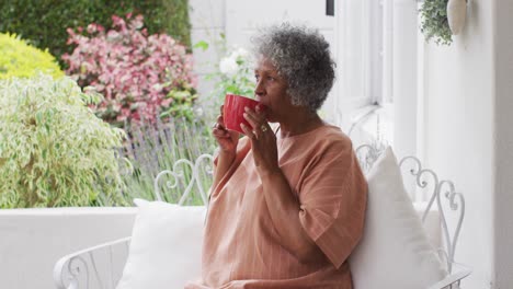 Ältere-Afroamerikanische-Frau-Trinkt-Kaffee,-Während-Sie-Auf-Der-Veranda-Des-Hauses-Sitzt