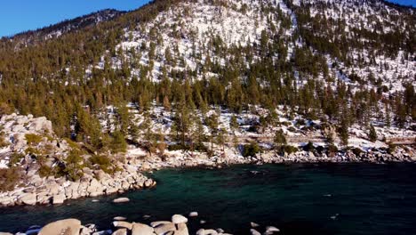 Aerial-drone-shot,-flying-over-the-lake-and-the-woods-towards-the-snowy-mountain-wall-in-Lake-Tahoe,-Nevada-California