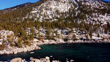 Toma-Aérea-De-Drones,-Volando-Sobre-El-Lago-Y-El-Bosque-Hacia-La-Pared-Nevada-De-La-Montaña-En-El-Lago-Tahoe,-Nevada-california