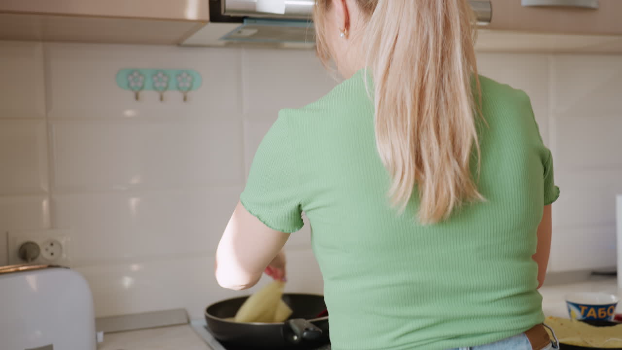 Blonde woman in green shirt seen from behind as she prepares breakfast in bright tiled kitchen, turning pancake on stove under cabinets with light