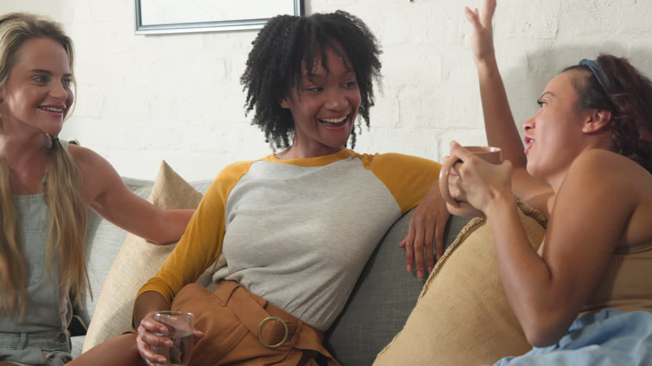 Laughing together, diverse female friends relaxing on couch, enjoying drinks indoors
