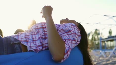 Junge-Frau-Liegt-Auf-Einem-Sessel-Und-Hört-Einem-Mann-Zu,-Der-In-Einer-Gruppe-Von-Freunden-An-Einem-Sommerabend-Bei-Sonnenuntergang-Am-Strand-Sitzt-Und-Gitarre-Spielt.-Zeitlupenaufnahme