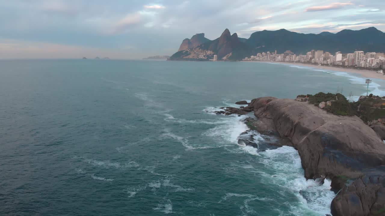 movimiento aéreo hacia atrás que muestra las olas chocando contra las rocas del acantilado de arpoador en primer plano y el paisaje urbano más amplio de la playa de ipanema y río de janeiro en el fondo