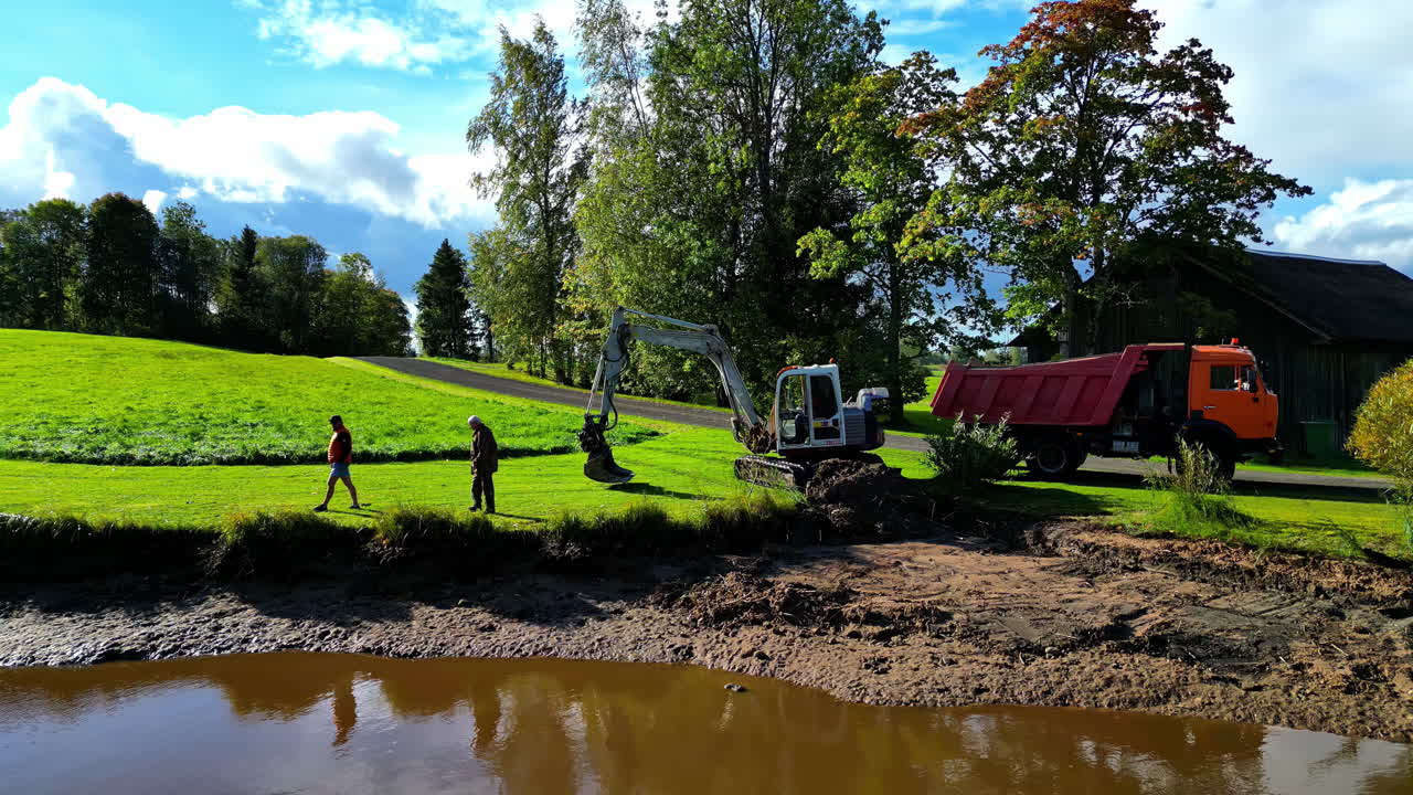 An excavator works on clearing a riverbank near a rural house and a truck, with workers overseeing the process in Latvia