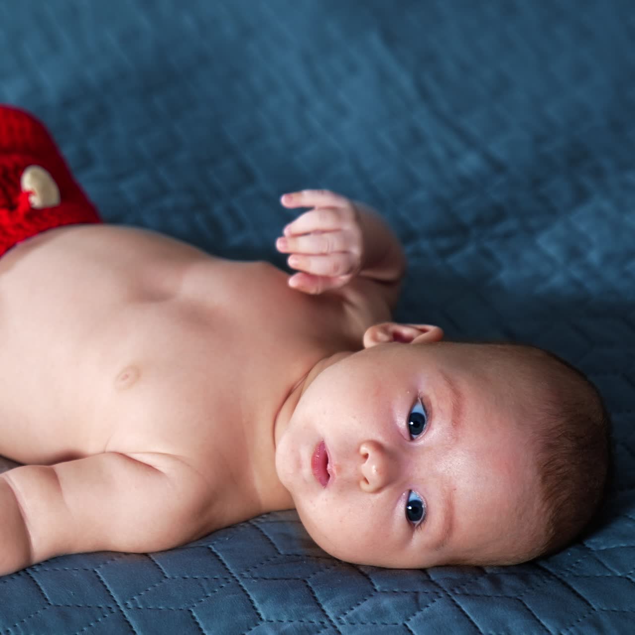 Blue-eyed Caucasian baby boy lying on bed wearing red shorts. Cute kid with naked belly tossing feet. Grey backdrop