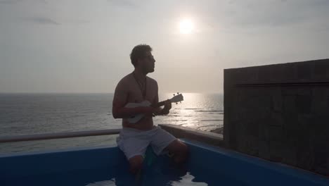 a man singing and playing a ukulele guitar sitting in a pool on the terrace of a beach house with the background of the sea and the sunset in 4k