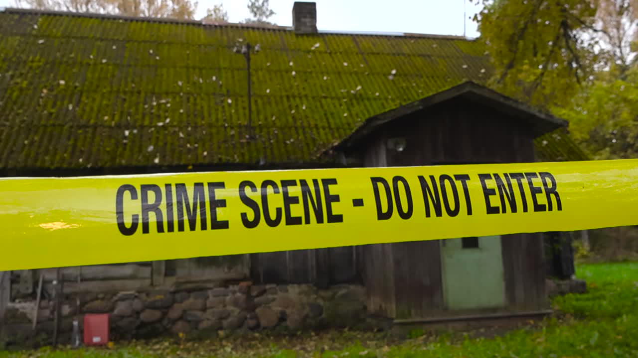 Yellow colored police crime scene forensic investigation tape ribbon in front of an old wooden traditional farm house or barn shed during a autumn day with fallen yellow leaves on the ground, bokeh