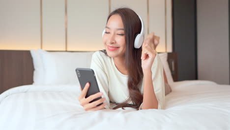 beautiful happy smiling thai woman lying on bed with headphones listening to music while holding mobile phone in hotel room