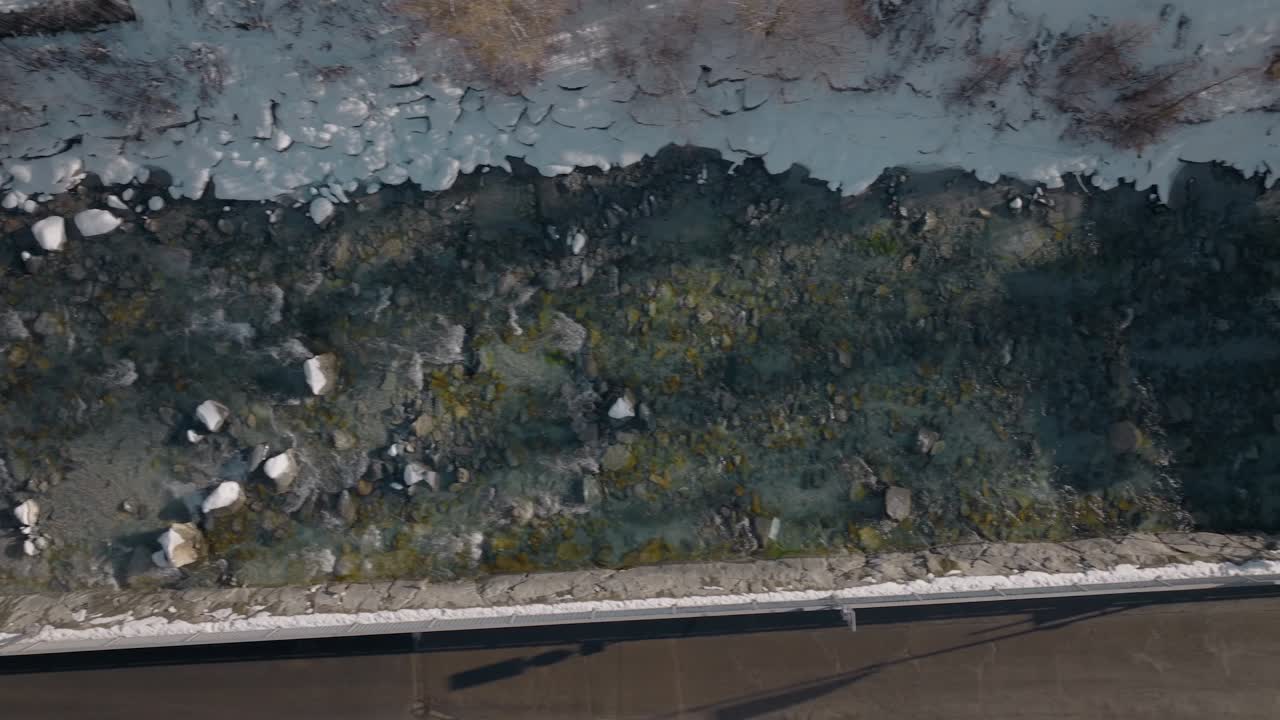 Aerial top down shot of cute small shallow river in Swiss village flowing alongside rocks and under bridge on beautiful sunny winter day with snow on side of river bank