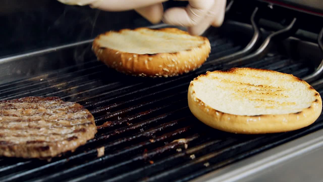 Frying cutlet and buns for hamburger. Chef is turning over roasted halves of bread for burger on hot grill. Fast food preparation