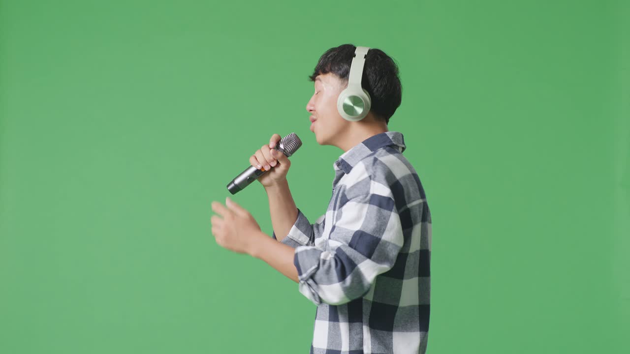젊은 아시아 십대 소년이 헤드폰을 착용하고 마이크를 들고 녹색 화면 배경에서 노래하는 측면 시각