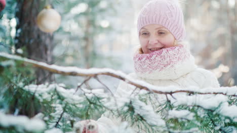 A-Middle-Aged-Woman-In-A-Knitted-Warm-Hat-Decorates-A-New-Year-Tree-In-A-Snow-Covered-Forest