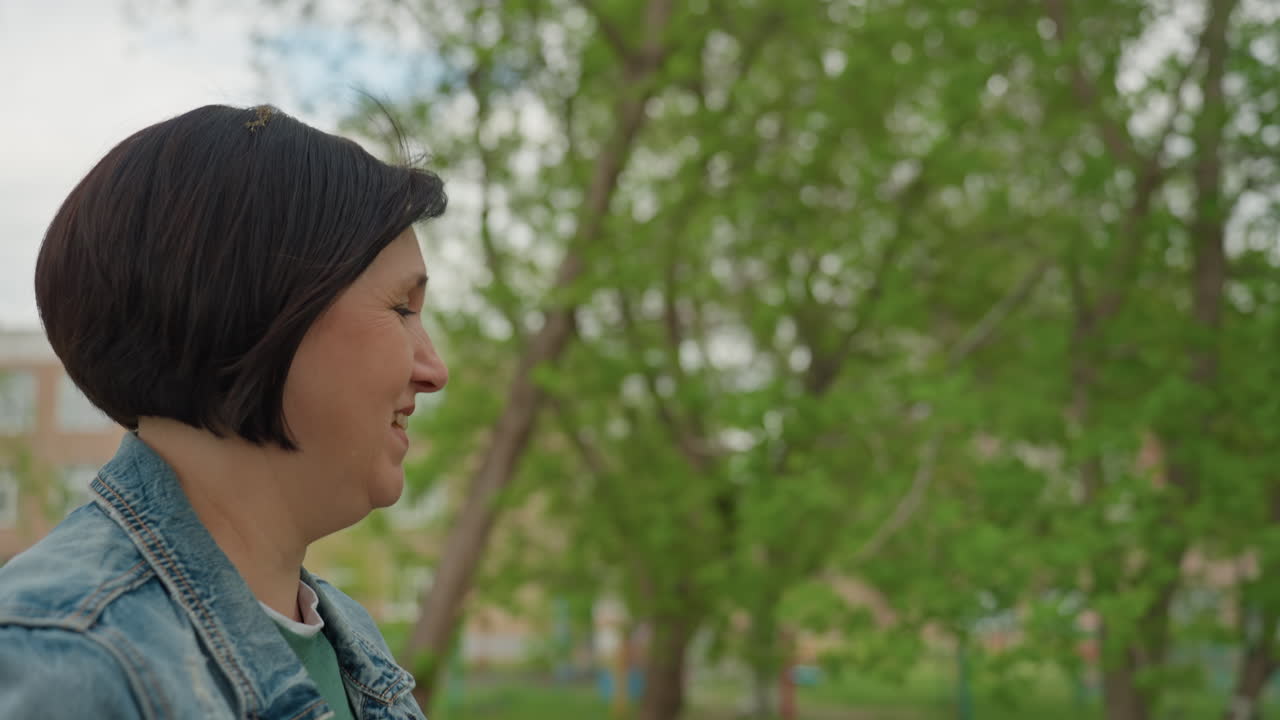 Caucasian Woman Profile In Park Wearing Denim Jacket And Short Bob Haircut, Pauses On Walking Path With Thoughtful Gaze Toward Trees And Soft Urban Background, Quiet Reflective Mood Suggesting