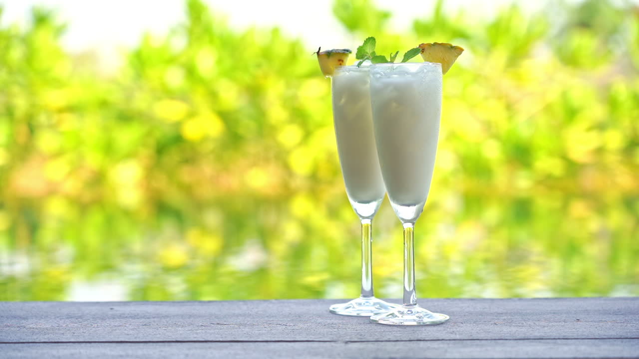 Two tropical drinks sit on the edge of a wooden deck overlooking the water and green and yellow backdrop