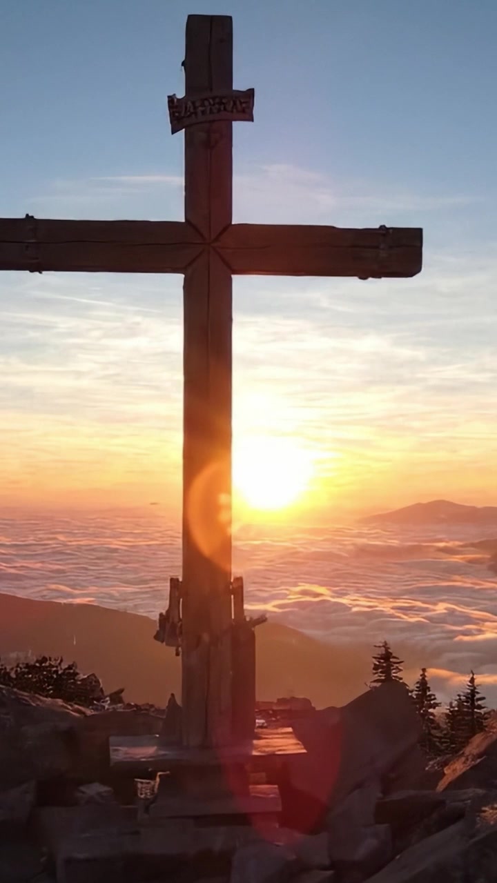 Uno splendido tramonto sulle montagne incorniciato da una croce di legno, che illumina il cielo con colori caldi e proietta ombre sul terreno roccioso sottostante.
