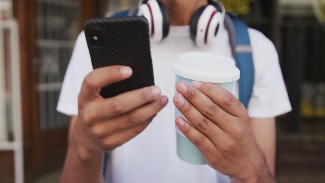 sección media de un hombre de raza mixta bebiendo café y usando un teléfono inteligente en la calle