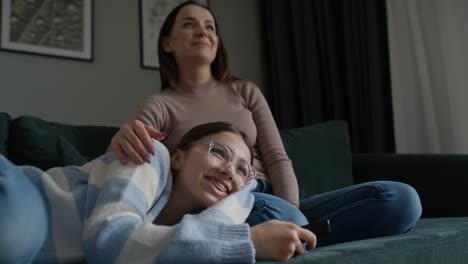 adolescente caucásico manteniendo la cabeza sobre las piernas de la madre mientras se acuesta en el sofá y mira la televisión