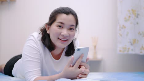 Mujer-Asiática-Tailandesa-Que-Mira-Y-Toca-El-Teléfono-Inteligente,-Relájese,-Disfrute-Y-Sonría-Con-Las-Redes-Sociales-En-Línea-En-El-Dormitorio