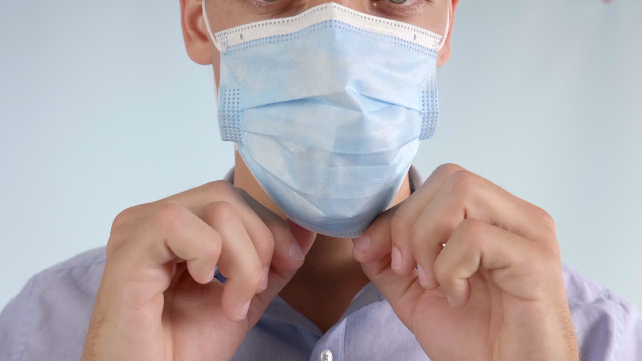 un hombre adulto feliz con ojos color avellana que usa una camisa abotonada se pone una máscara quirúrgica en un fondo gris para protegerse del covid-19 - coronavirus, directamente en una toma de trípode de cerca