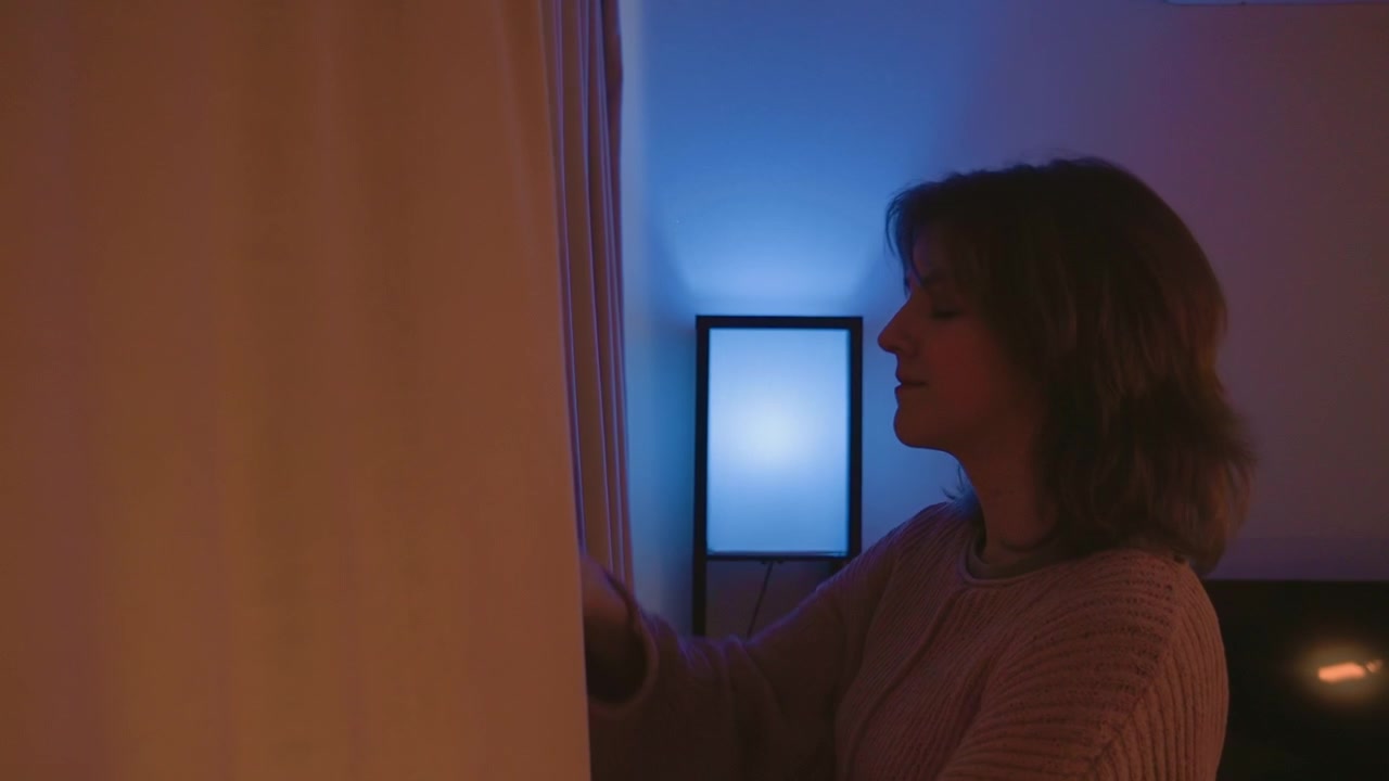 Kvinde i profil glatter et gardin med løftet hånd, mens hun kigger mod et lyst blåt vægpanel, hvilket skaber en rolig, moderne indendørsscene med blød kontrast, en afslappet stemning og subtil bevægelse.