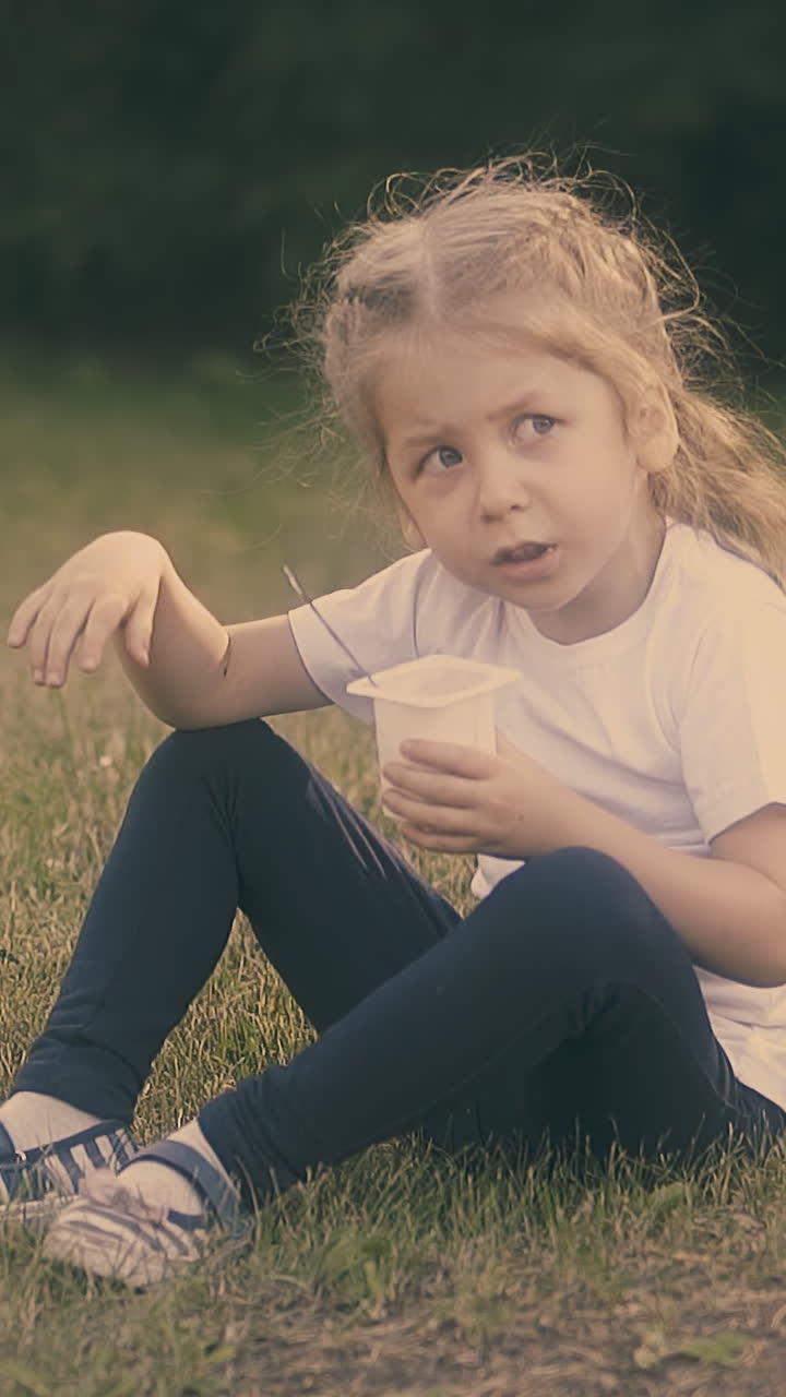 nice little blond girl in white t-shirt and black pants eats tasty yogurt sitting on green grass close view slow motion