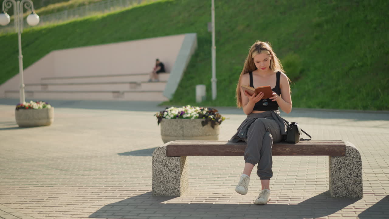 웃는 듯이 벤치에 앉아서 책을 읽고, 다리를 고, 태양이 그녀를 비추고, 배경에는 멀리 있는 사람들, 조명 기둥, 그리고 울창한 초록색이 있습니다.