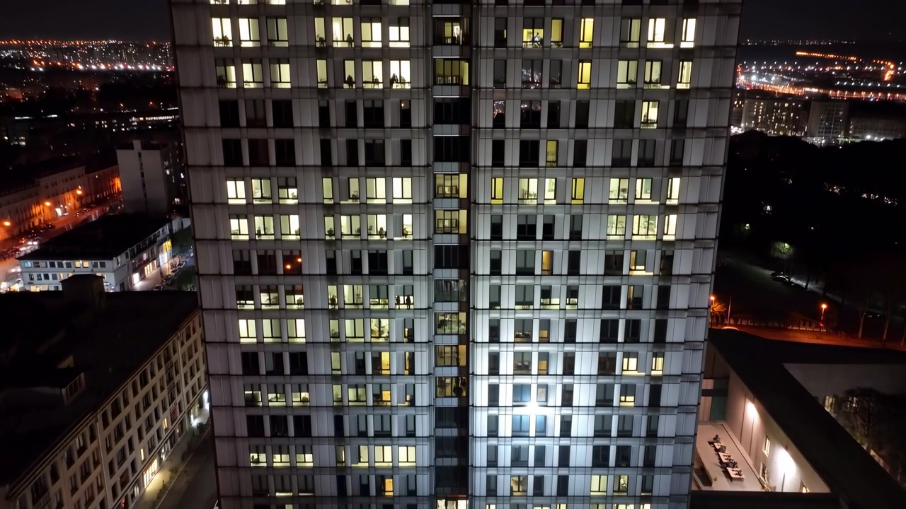 Veduta aerea notturna cattura la facciata illuminata di un grattacielo con file di finestre luminose, dintorni scuri e luci della città distanti, creando una composizione urbana dettagliata da una prospettiva elevata.