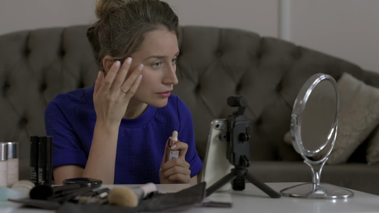 Woman in a blue T-shirt filming herself while doing a make-up tutorial at home