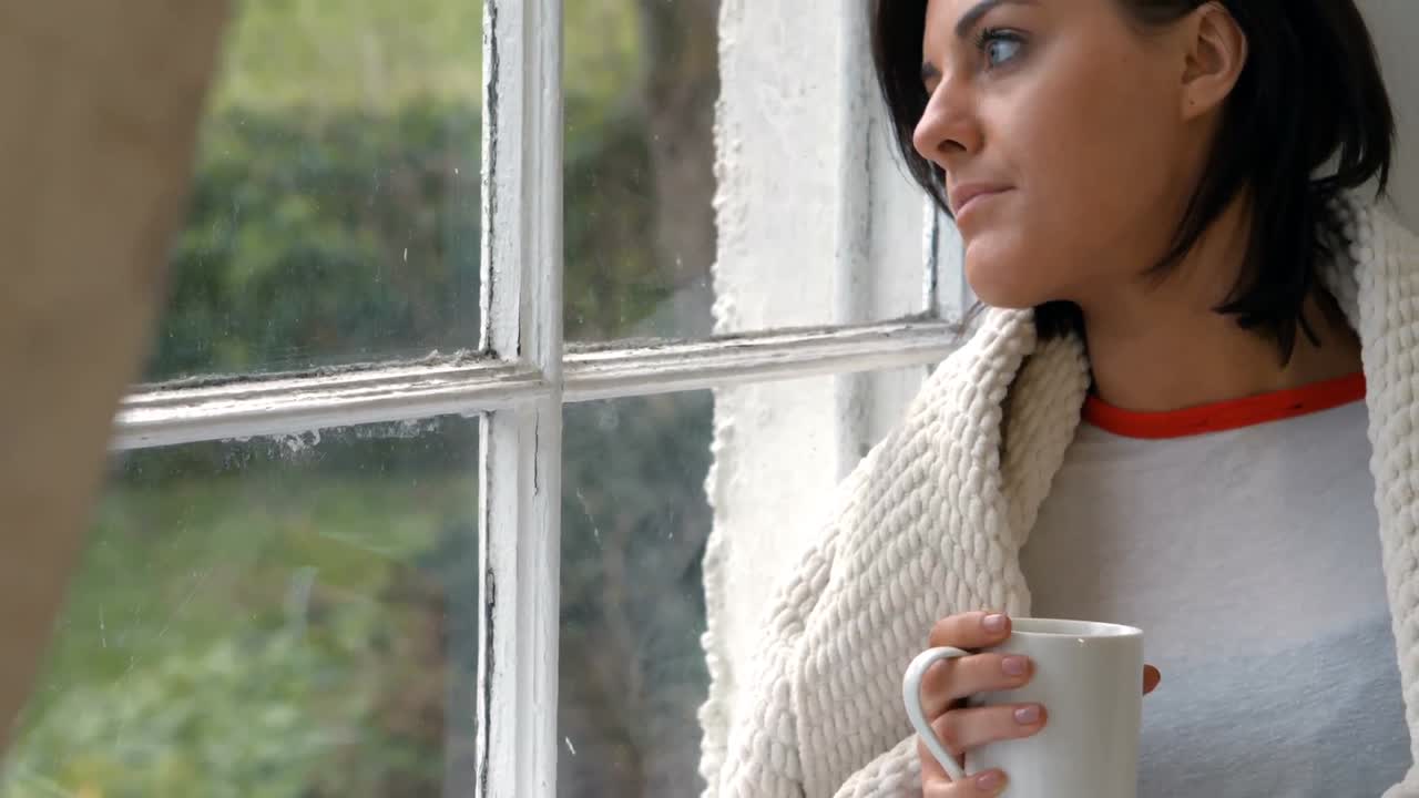 Donna pensierosa seduta sul davanzale della finestra che beve una tazza di caffè