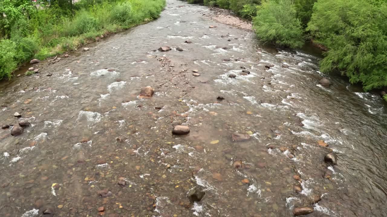 Clear shallow stream of the Talvera River winding over smooth stones and gravel, bordered by lush green vegetation along the Lungotalvera walking paths in the city of Bolzano