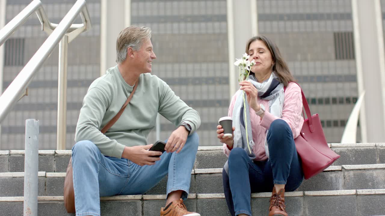 Pareja sénior sonriendo y charlando en escaleras urbanas sosteniendo margaritas, inhalando su aroma y después bebiendo café