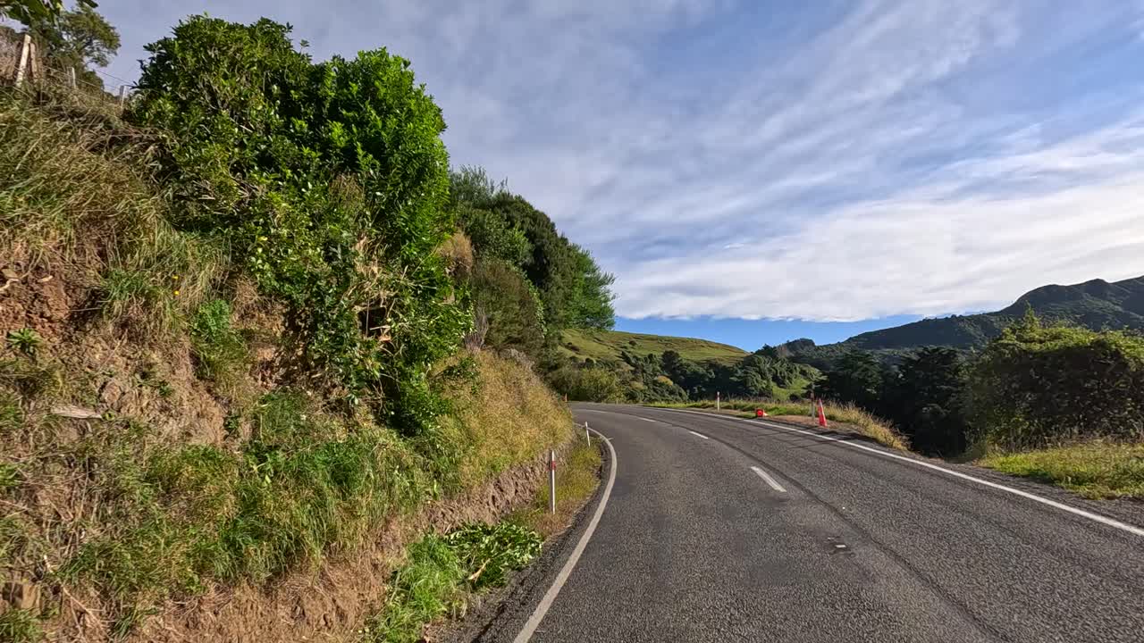 A serene drive along a winding rural road in Akaroa, New Zealand, showcasing lush greenery and expansive landscapes under a bright sky