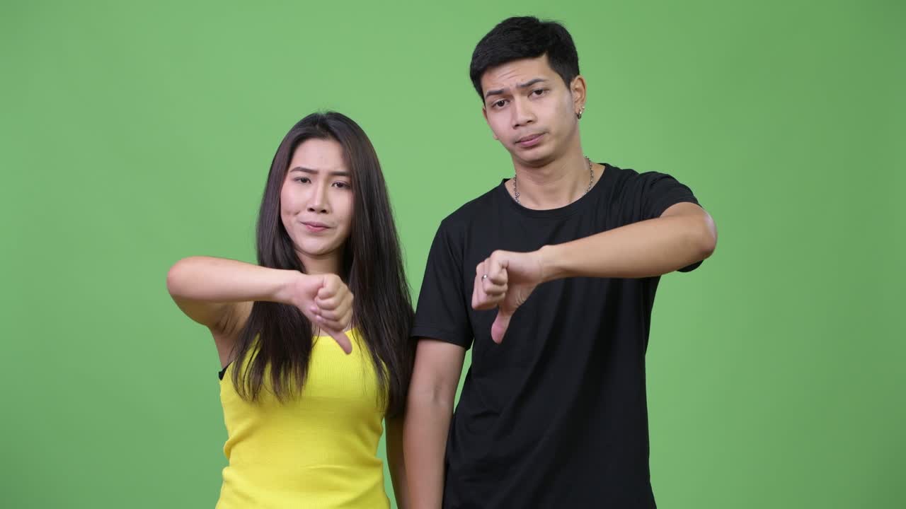 Young sad Asian couple giving thumbs down together