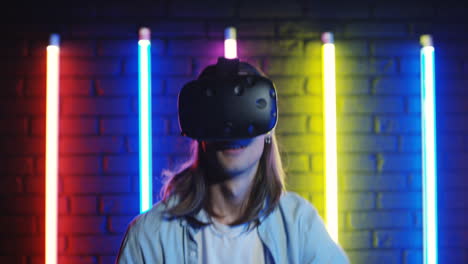 Vista-De-Cerca-De-Un-Joven-Que-Mira-La-Cámara,-Luego-Se-Pone-Gafas-Vr-Y-Comienza-A-Jugar-Un-Juego-Virtual-En-Una-Habitación-Con-Coloridas-Lámparas-De-Neón-En-La-Pared