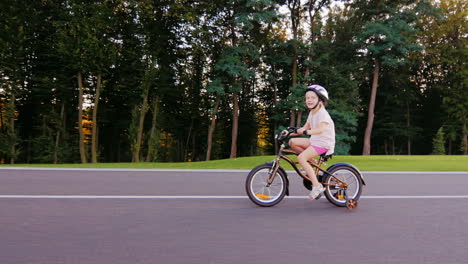 Girl-Five-Years-In-A-Helmet-Riding-A-Bike-In-The-Well-Kept-Park-On-A-Background-Of-Trees